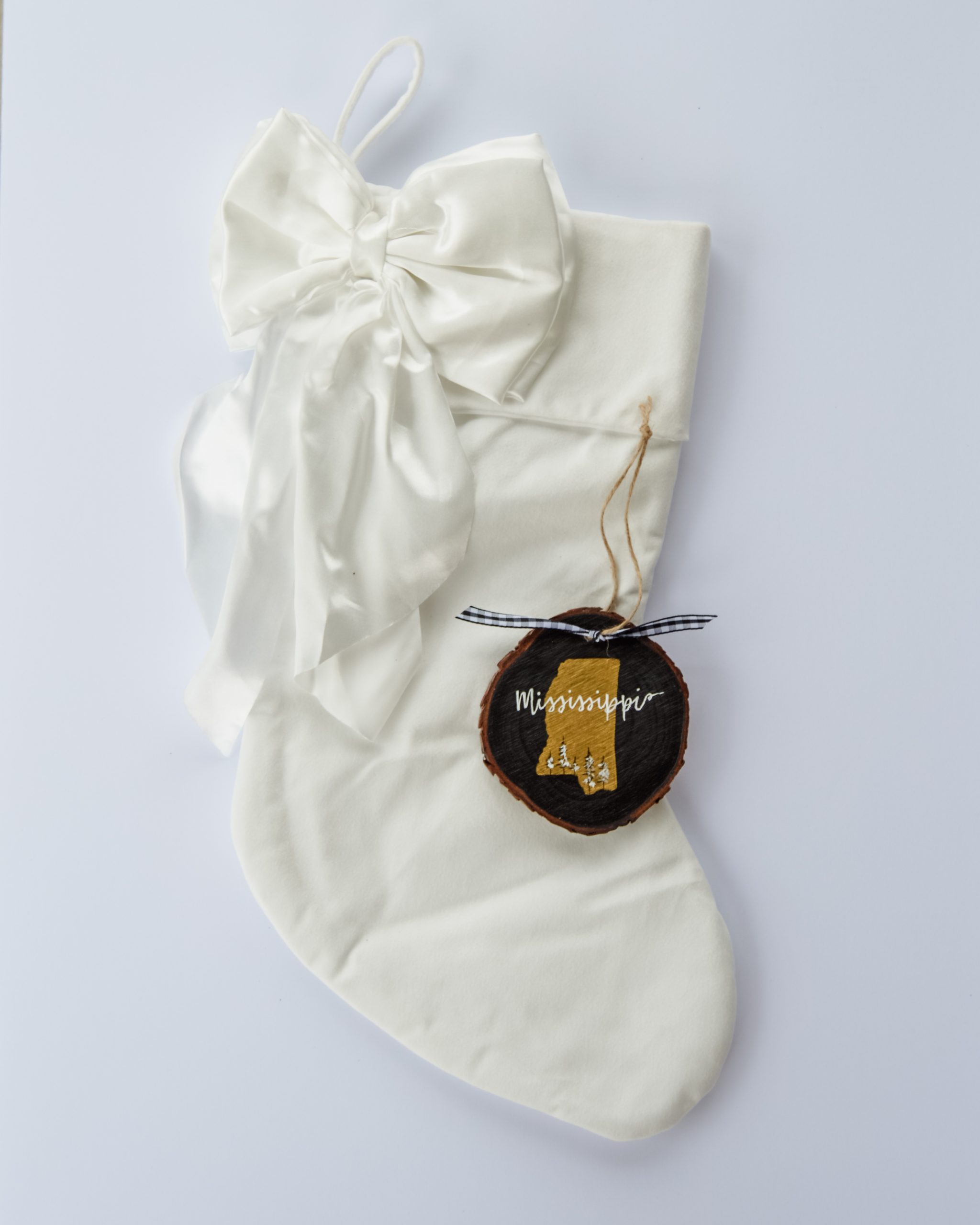 Hand-painted wood slice ornament of Mississippi with a gold state silhouette, snow-covered trees, and the state name hand-lettered in white, tied with white buffalo plaid ribbon on a white velvet Christmas stocking