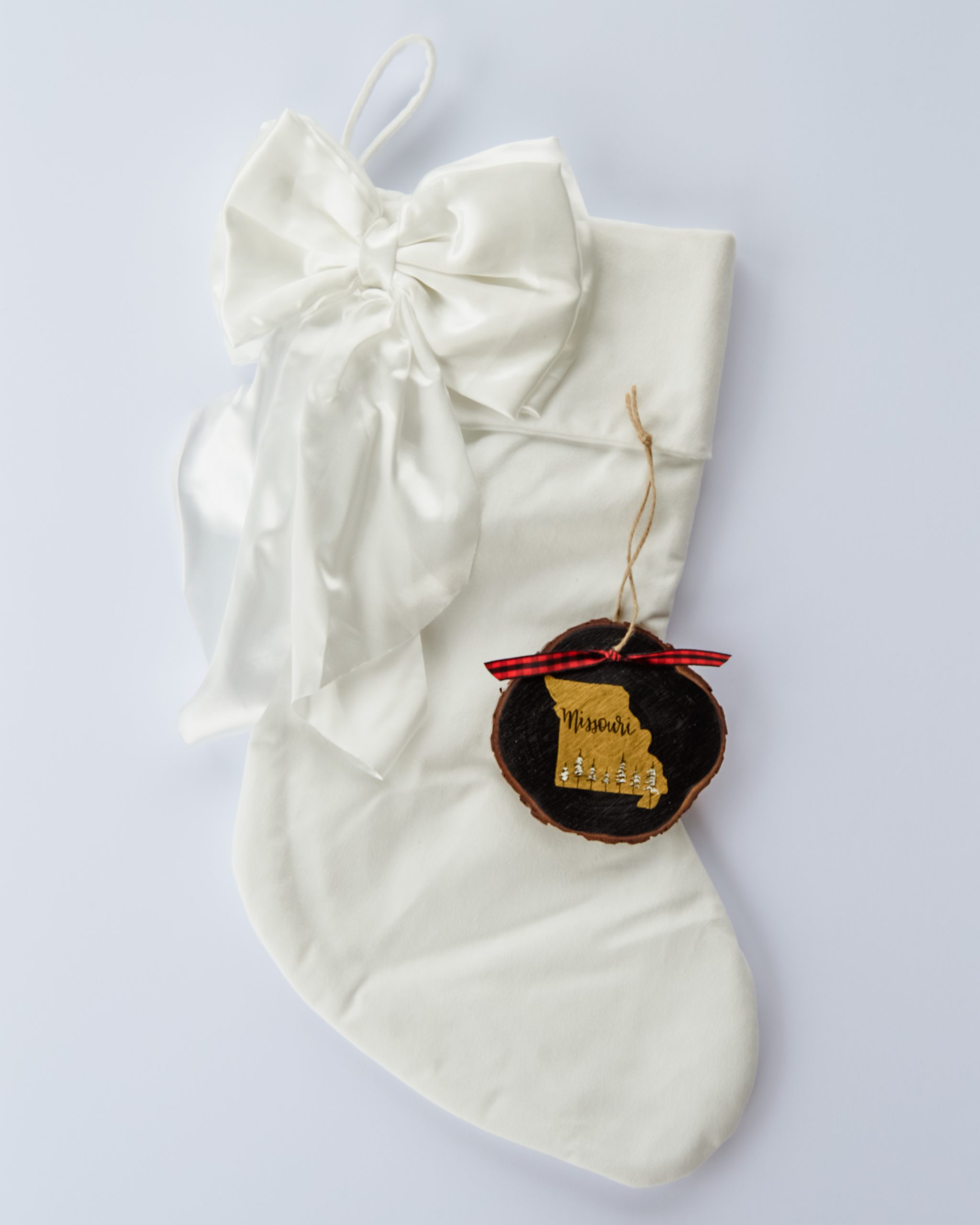 Hand-painted wood slice ornament of Missouri with a gold state silhouette, snow-covered trees, and the state name hand-lettered in black, tied with red buffalo plaid ribbon on a white velvet Christmas stocking