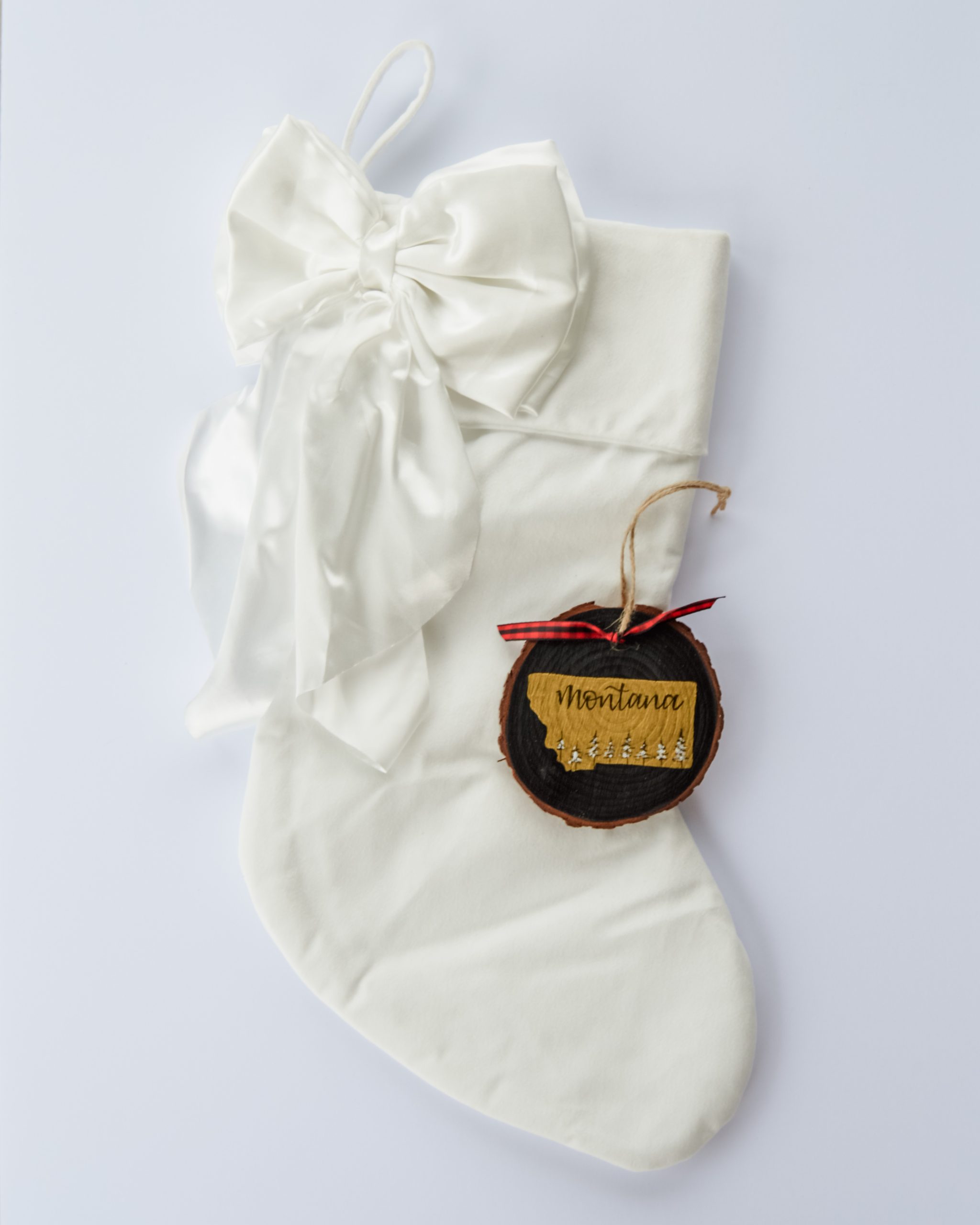 Hand-painted wood slice ornament of Montana with a gold state silhouette, snow-covered trees, and the state name hand-lettered in black, tied with red buffalo plaid ribbon on a white velvet Christmas stocking