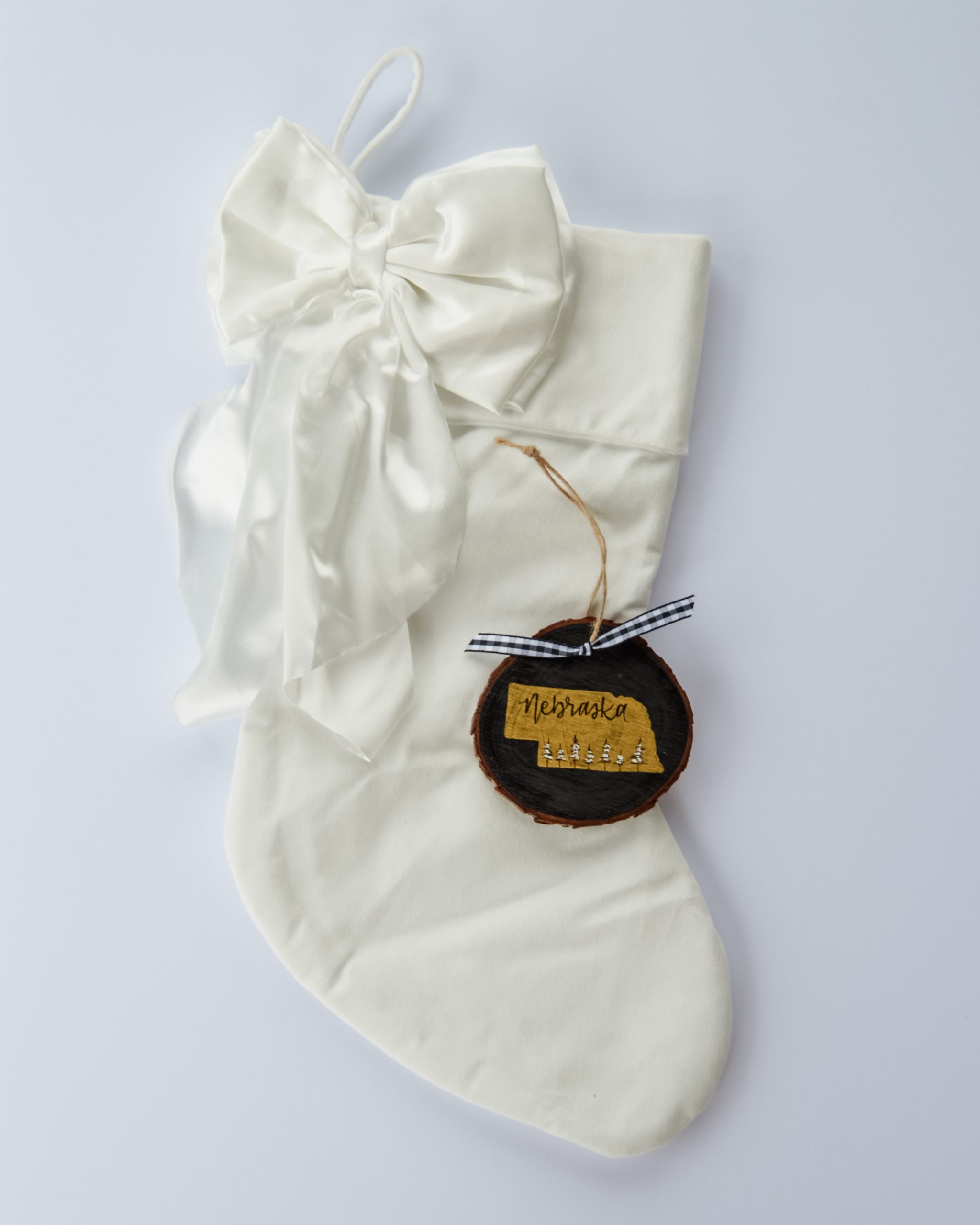 Hand-painted wood slice ornament of Nebraska with a gold state silhouette, snow-covered trees, and the state name hand-lettered in black, tied with white buffalo plaid ribbon on a white velvet Christmas stocking