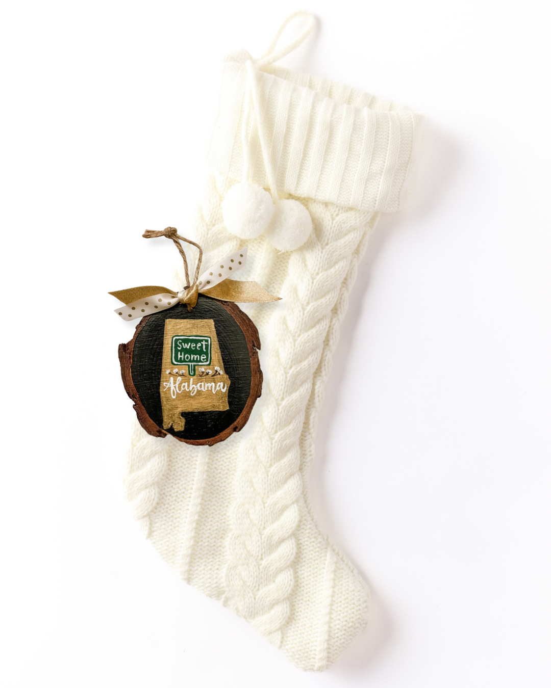 Hand-painted wood slice ornament with a gold Alabama silhouette, a green "Sweet Home" road sign, white cotton bolls, white hand-lettered "Alabama" on black, and gold and polka dot ribbon on a white knit stocking