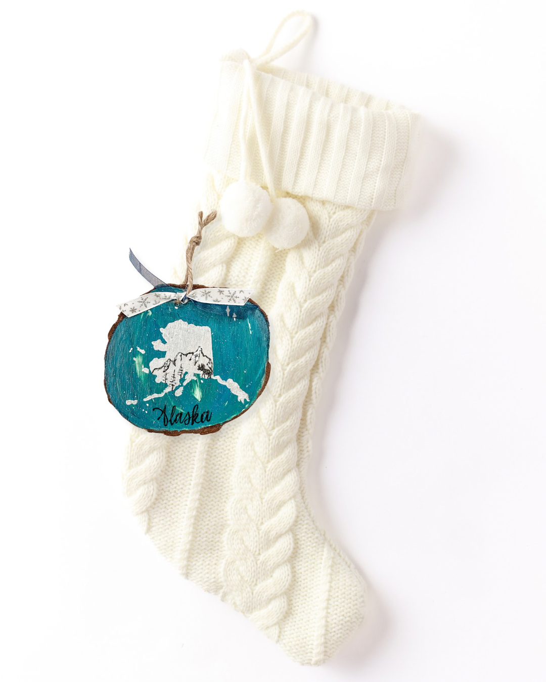 Hand-painted wood slice ornament with a white Alaska silhouette with a sketched mountain, trees, and moose on a teal Northern Lights background, hand-lettered "Alaska," and snowflake ribbon on a white knit stocking