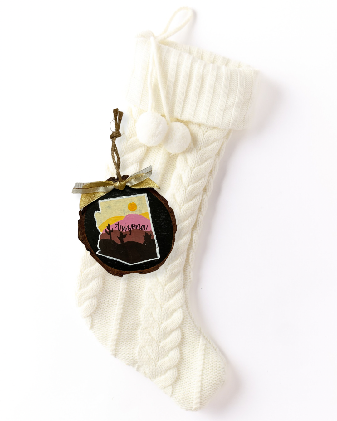 Hand-painted wood slice ornament with an Arizona silhouette showing a desert sunset with pink and brown mountains and saguaro cacti on black, hand-lettered "Arizona," and gold ribbon on a white knit stocking