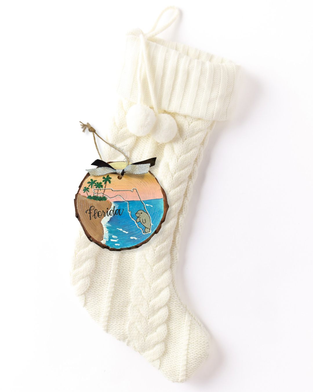 Hand-painted wood slice ornament with a Florida outline over a beach scene with palm trees, turquoise water, a sunset sky, and a manatee, hand-lettered "Florida," and black and silver ribbon on a white knit stocking