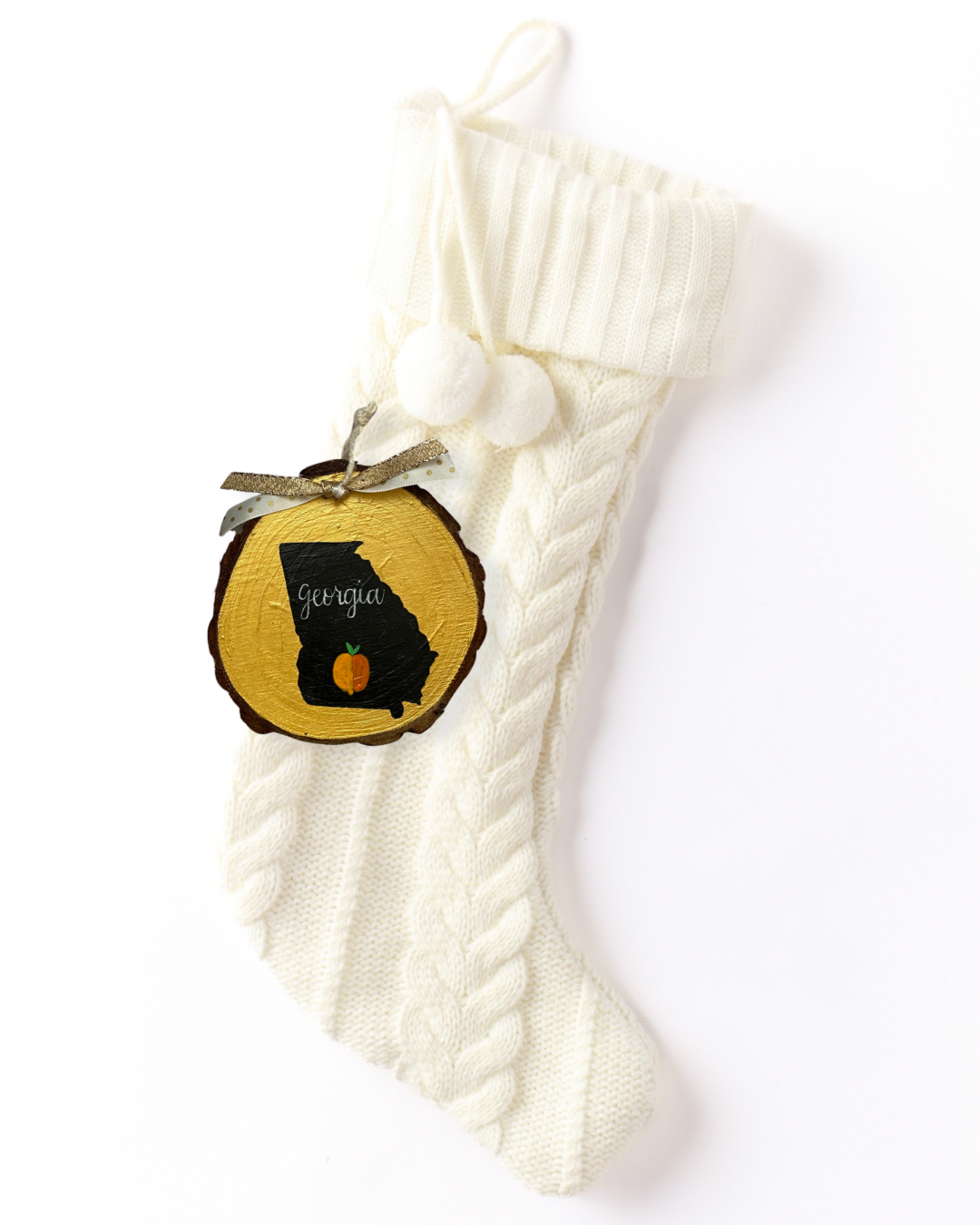 Hand-painted wood slice ornament with a black Georgia silhouette on gold, a small orange peach, white hand-lettered "Georgia," and gold glitter ribbon on a white knit stocking