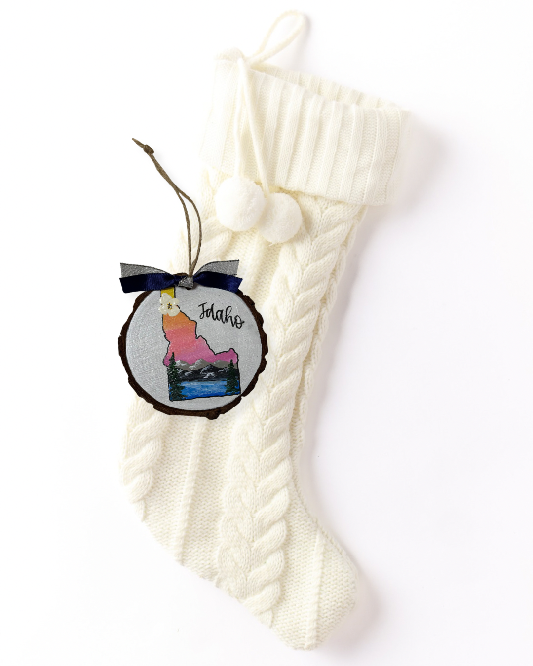 Hand-painted wood slice ornament with an Idaho silhouette filled with a pink sunset, mountains, evergreens, and a blue lake, hand-lettered "Idaho," and navy ribbon on a white knit stocking