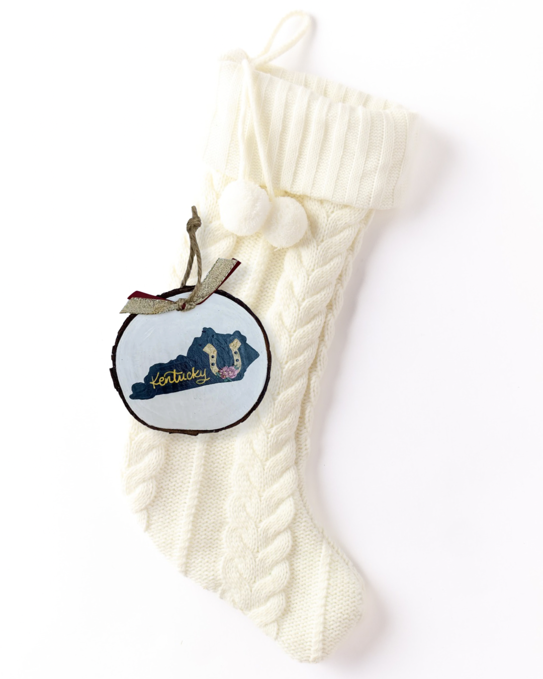 Hand-painted wood slice ornament with a navy Kentucky silhouette, a gold horseshoe with a pink rose, gold hand-lettered "Kentucky," and gold and burgundy ribbon on a white knit stocking