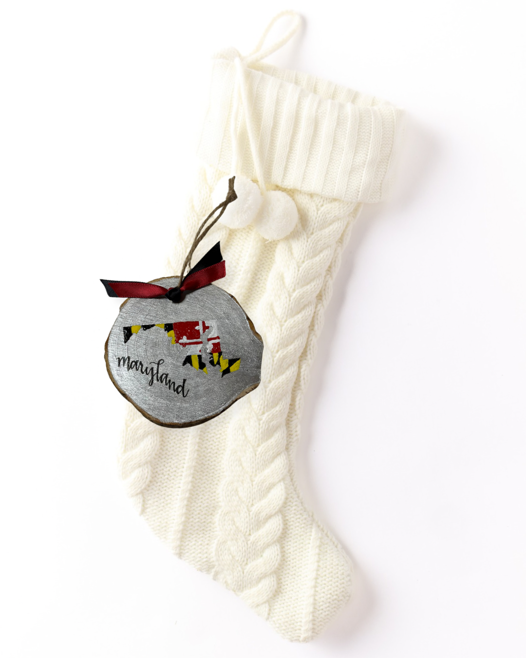 Hand-painted wood slice ornament with a Maryland silhouette in the state flag pattern on a silver background, hand-lettered "Maryland," and red and black ribbon on a white knit stocking