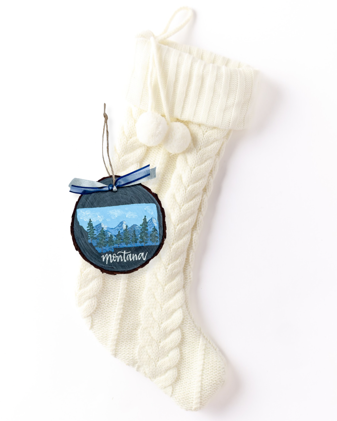 Hand-painted wood slice ornament with a Montana silhouette filled with blue snowy mountains and evergreen trees on slate gray, white hand-lettered "Montana," and blue ribbon on a white knit stocking