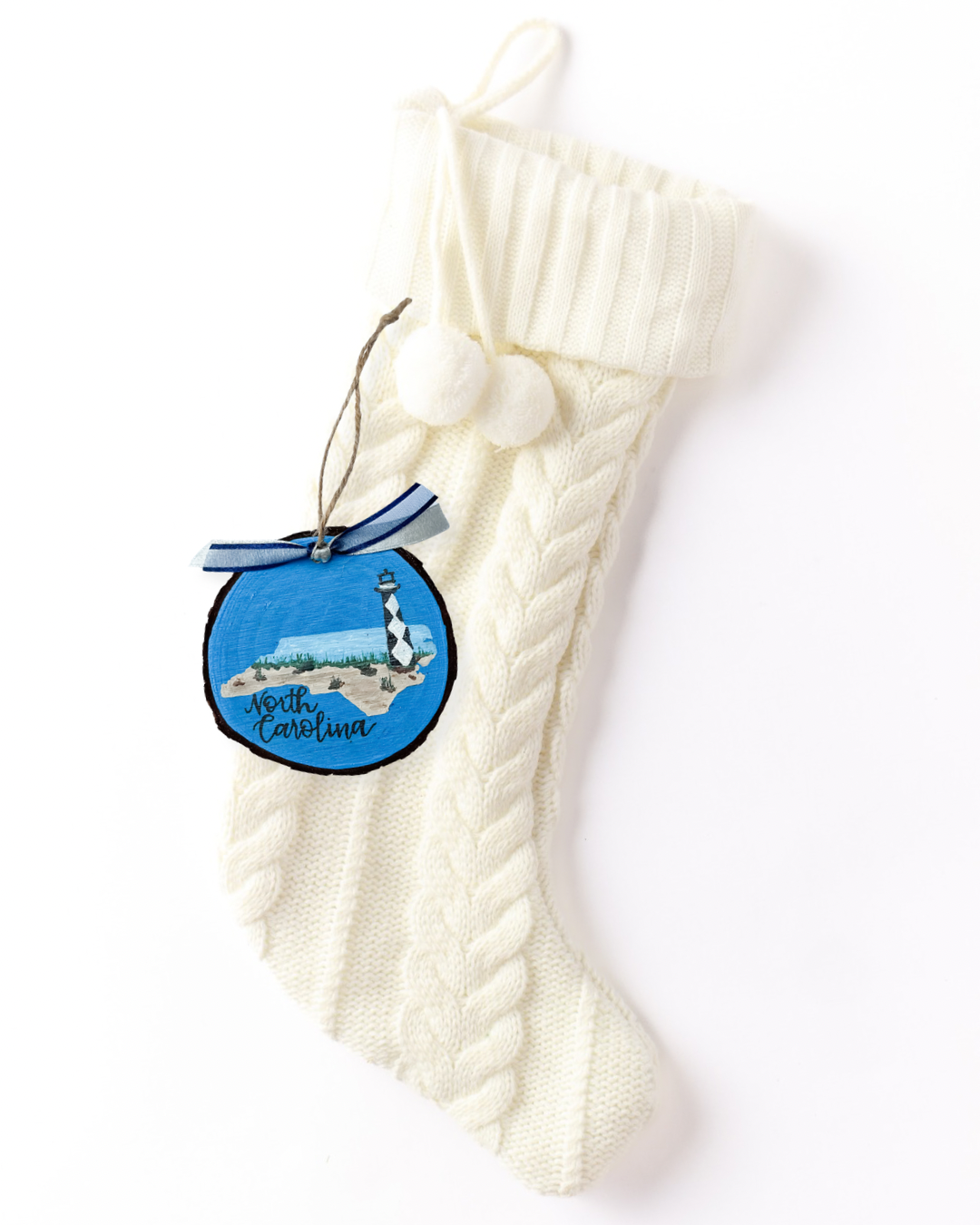 Hand-painted wood slice ornament with a North Carolina silhouette showing a beach, dune grass, and black and white diamond lighthouse on blue, hand-lettered "North Carolina," and blue ribbon on a white knit stocking