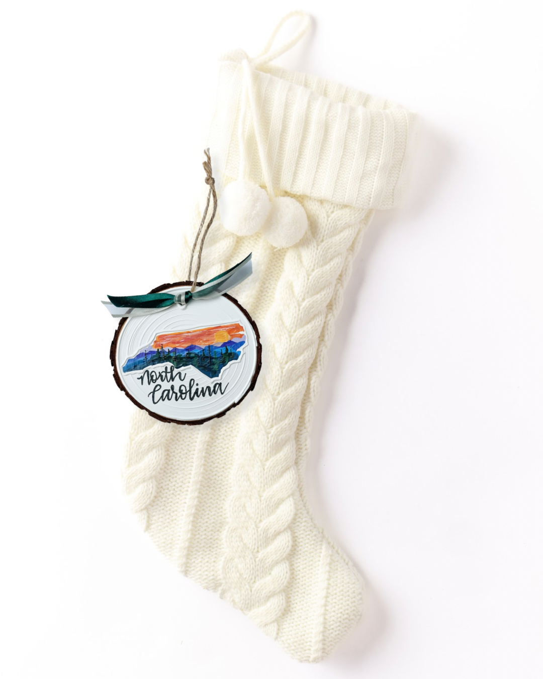 Hand-painted wood slice ornament with a North Carolina silhouette filled with an orange sunset over blue mountains and evergreens, hand-lettered "North Carolina," and green ribbon on a white knit stocking