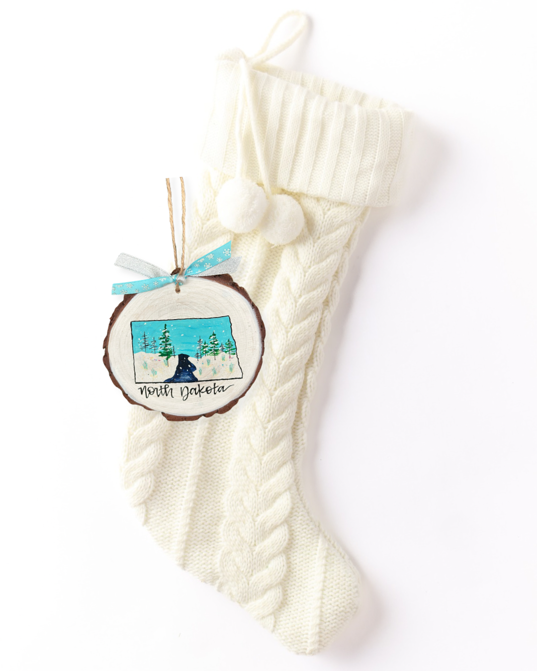 Hand-painted wood slice ornament with a North Dakota silhouette filled with a snowy evergreen forest and falling snow, hand-lettered "North Dakota," and blue snowflake ribbon on a white knit stocking