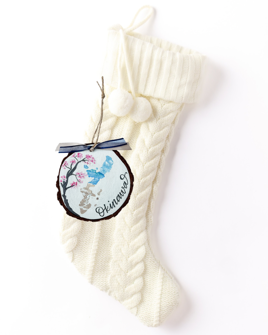 Hand-painted wood slice ornament with an Okinawa island silhouette in blue and sand tones, a pink cherry blossom tree, hand-lettered "Okinawa," and navy striped ribbon on a white knit stocking