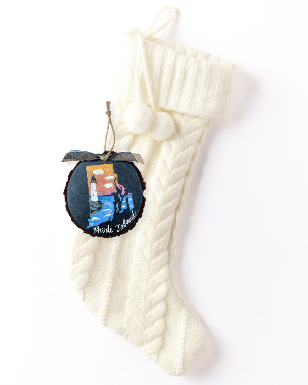 Hand-painted wood slice ornament with a Rhode Island silhouette featuring a white lighthouse, sunset sky, and ocean waves on navy, white hand-lettered "Rhode Island," and sheer ribbon on a white knit stocking