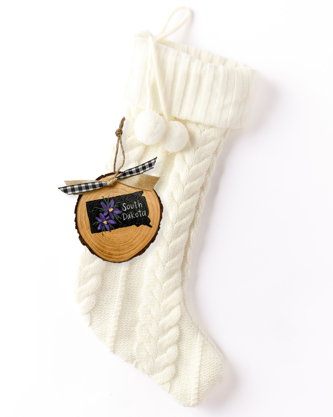 Hand-painted wood slice ornament with a black South Dakota silhouette, purple pasque flowers, white hand-lettered "South Dakota," and black and white check and gold ribbon on a white knit stocking