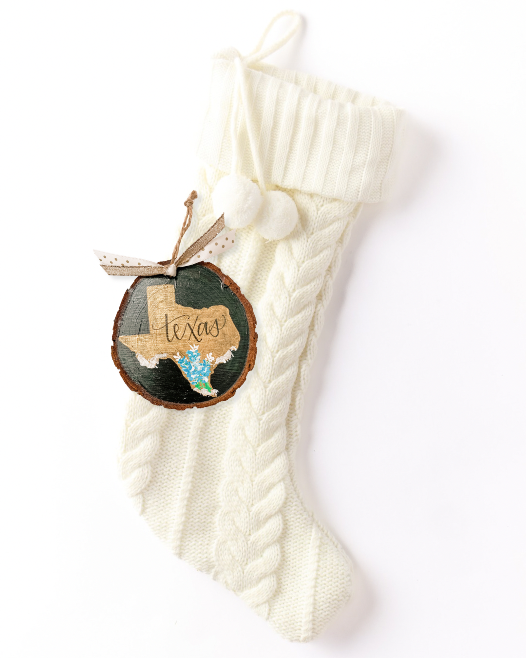 Hand-painted wood slice ornament with a gold Texas silhouette, blue bluebonnet flowers, hand-lettered "Texas," on a dark background with gold and polka dot ribbon on a white knit stocking