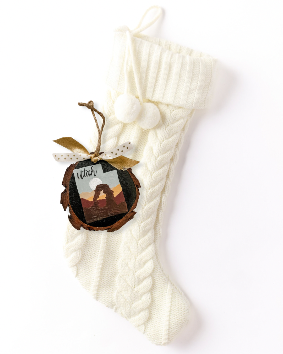 Hand-painted wood slice ornament with a Utah silhouette featuring Delicate Arch in red and orange desert tones, hand-lettered "Utah," and gold and polka dot ribbon on a white knit stocking
