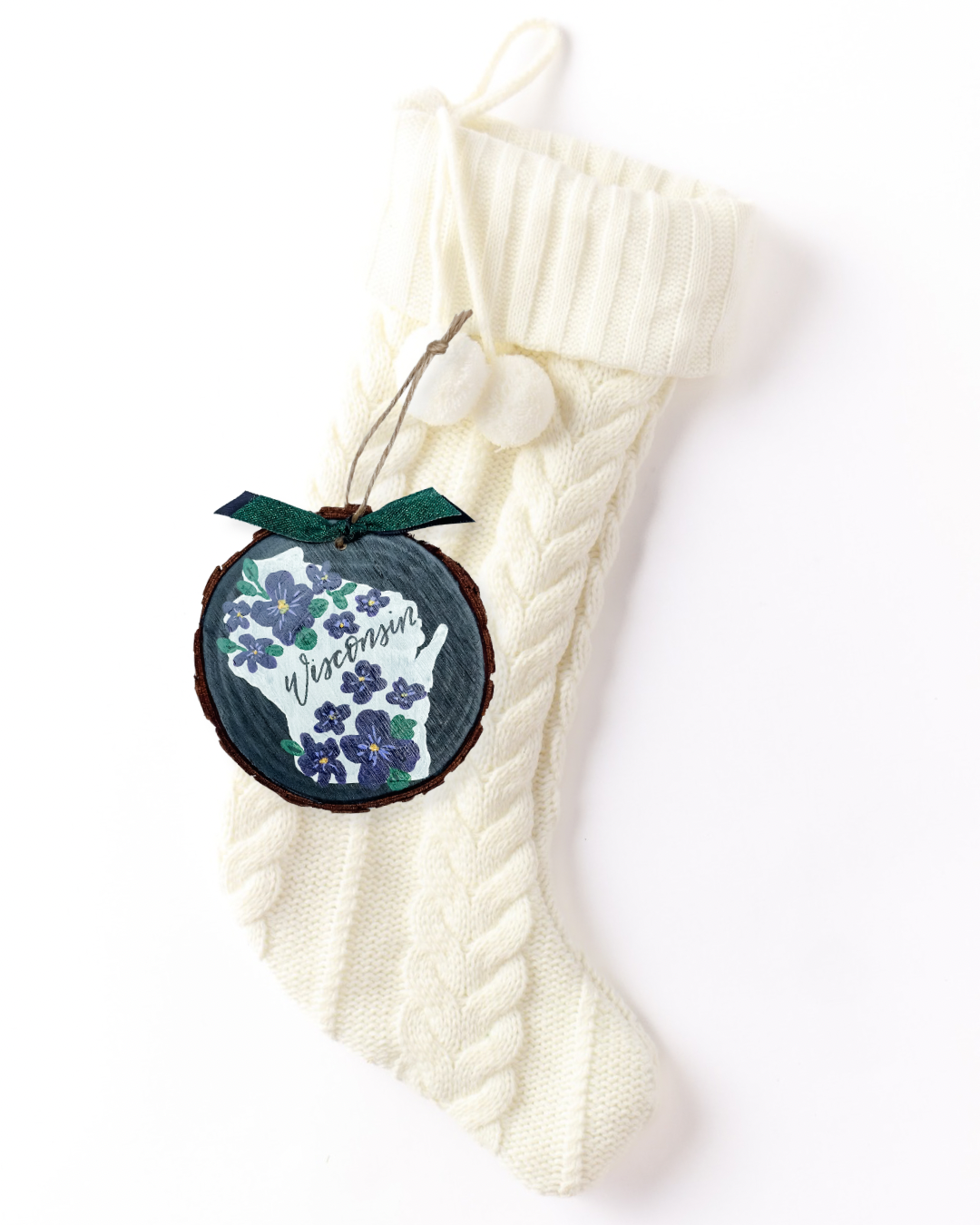 Hand-painted wood slice ornament with a white Wisconsin silhouette covered in purple wood violets on a charcoal background, hand-lettered "Wisconsin," and green ribbon on a white knit stocking