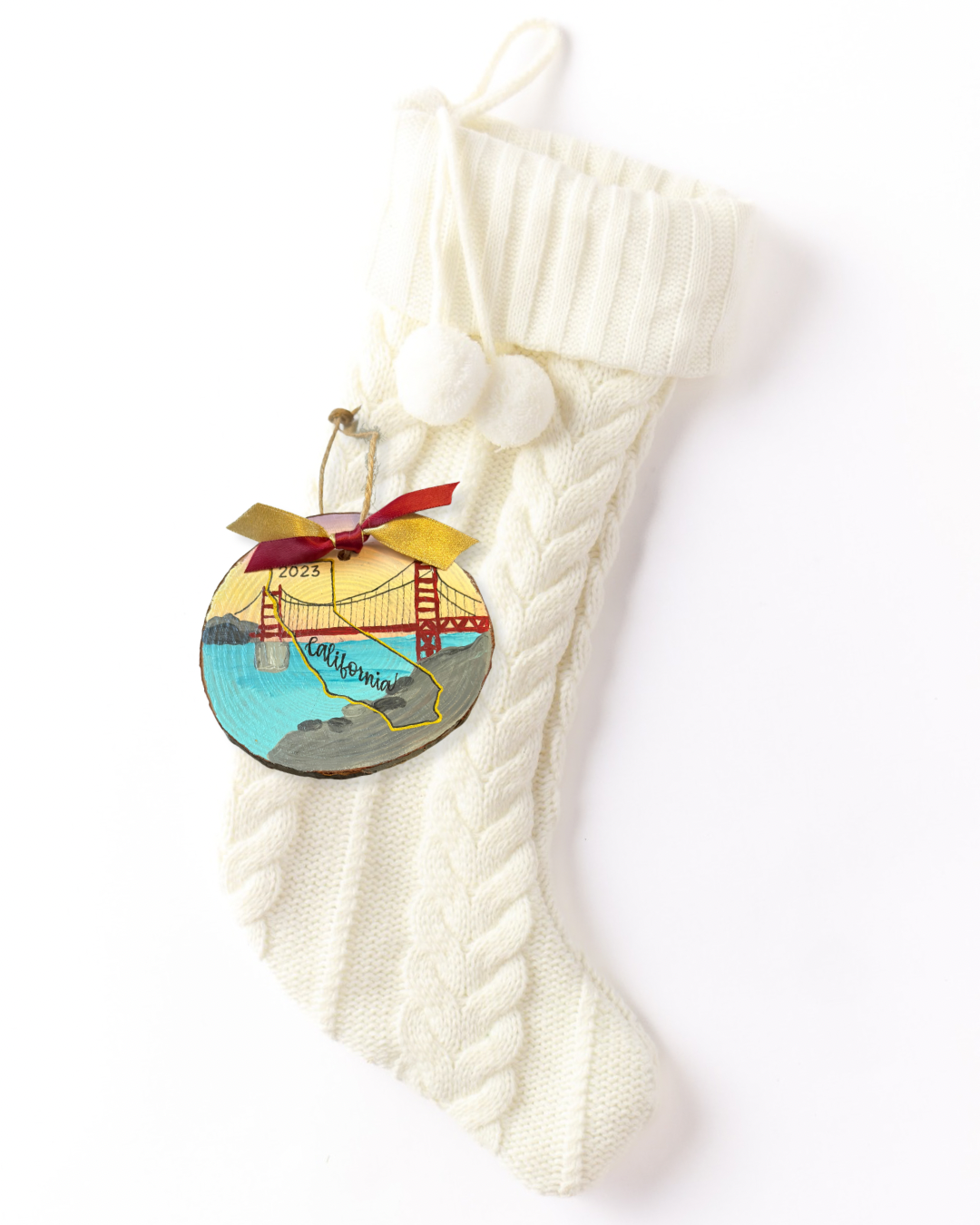 Hand-painted wood slice ornament with the Golden Gate Bridge at sunset, a gold outline of California, hand-lettered "California," and "2023," with red and gold ribbon on a white knit stocking