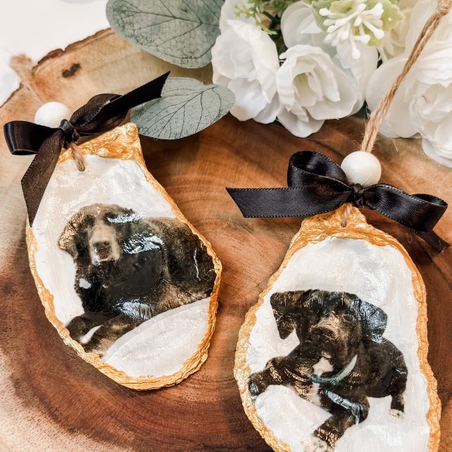 Two personalized oyster shell ornaments with gold edges and dog photos, tied with black satin bows, on a wood slice.