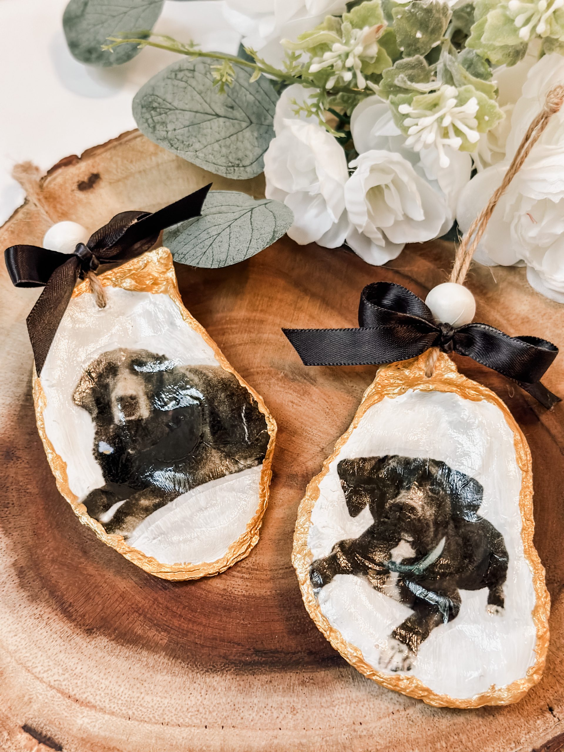 Two personalized oyster shell ornaments with gold edges and dog photos, tied with black satin bows, on a wood slice.