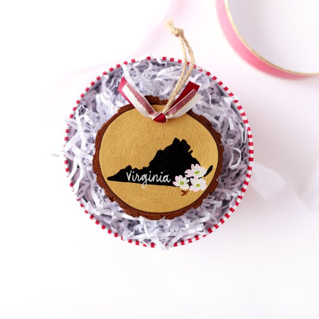 Hand-painted wood slice ornament with a black Virginia silhouette on gold, white dogwood blossoms, white hand-lettered "Virginia," and burgundy and white ribbon, resting on white crinkle paper in a red and white striped gift box with a pink lid nearby