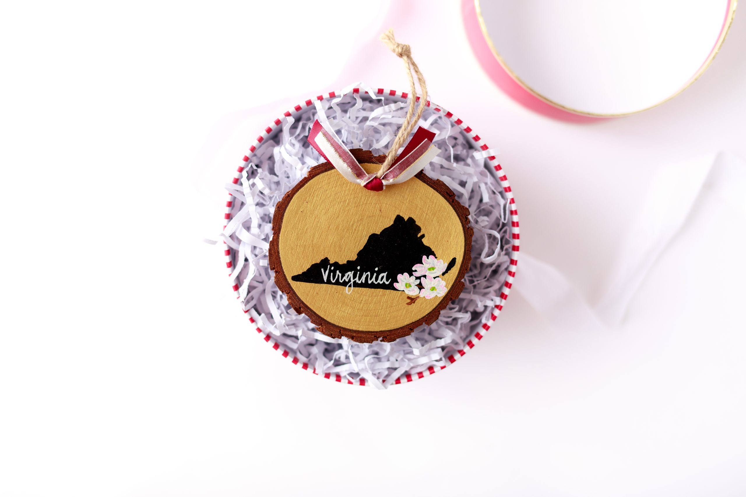 Hand-painted wood slice ornament with a black Virginia silhouette on gold, white dogwood blossoms, white hand-lettered "Virginia," and burgundy and white ribbon, resting on white crinkle paper in a red and white striped gift box with a pink lid nearby