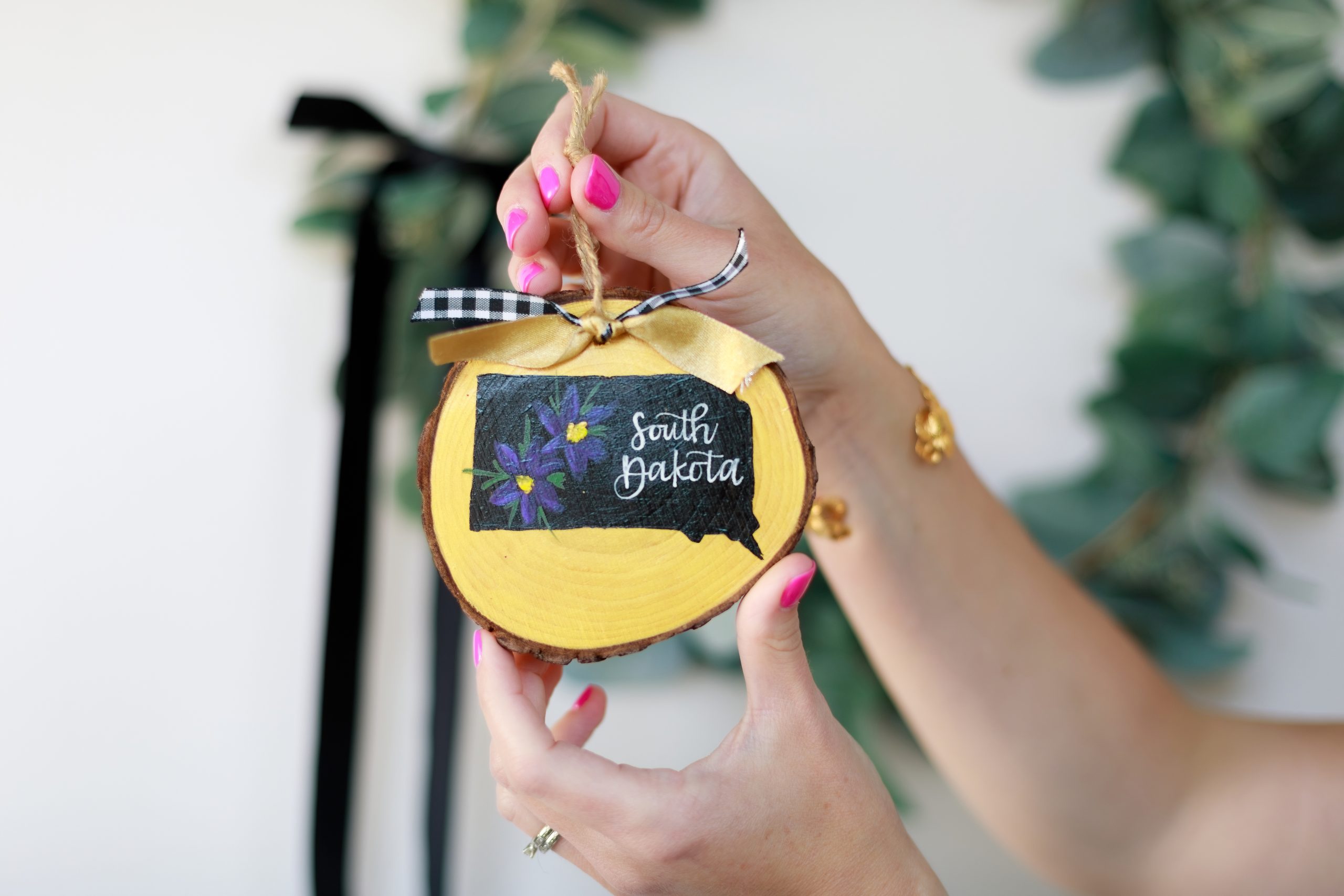 Hands with pink nails holding a hand-painted wood slice ornament by its jute twine, featuring a black South Dakota silhouette with purple pasque flowers, white hand-lettered "South Dakota," and black and white gingham and gold ribbon, with blurred greenery in the background