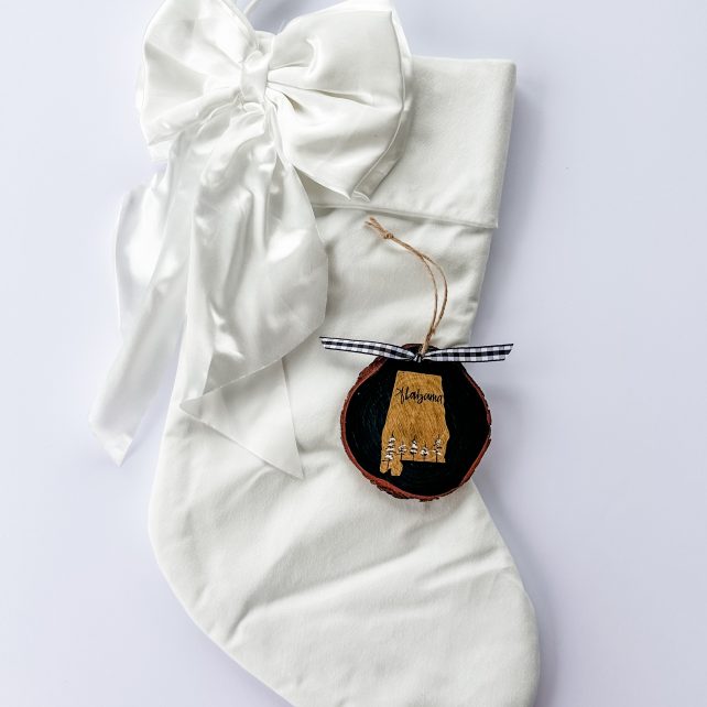 Hand-painted wood slice ornament of Alabama with a gold state silhouette, snow-covered trees, and the state name hand-lettered in black, tied with white buffalo plaid ribbon on a white velvet Christmas stocking