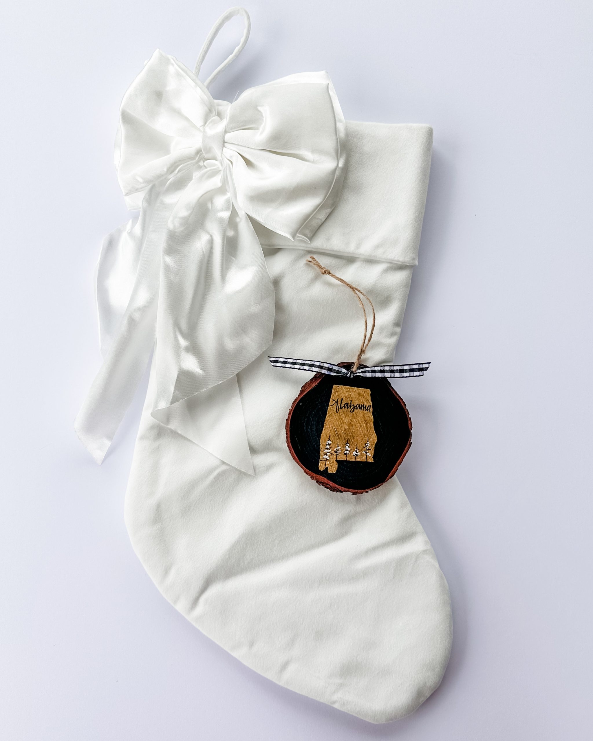 Hand-painted wood slice ornament of Alabama with a gold state silhouette, snow-covered trees, and the state name hand-lettered in black, tied with white buffalo plaid ribbon on a white velvet Christmas stocking